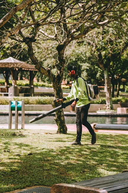A person wearing a light green jacket, black trousers, and red earmuffs operates a leaf blower to clear grass and fallen leaves from a well-maintained garden area. The garden features lush green grass, mature trees with thick, textured bark and leafy branches providing shade, and a calm pond visible in the background. The scene is set on a sunny day with soft natural lighting illuminating the garden, which is bordered by a paved pathway and a wooden bench. In the distance, a gazebo and some park furniture can be seen, indicating a public outdoor space. The individual appears focused on their gardening task, demonstrating proper lawn care and garden maintenance practices. The backdrop includes neatly trimmed foliage and a peaceful environment, exemplifying regular outdoor care typical of professional garden landscaping, as offered by Gardeners Haringey, who are experts in garden maintenance and landscaping work.